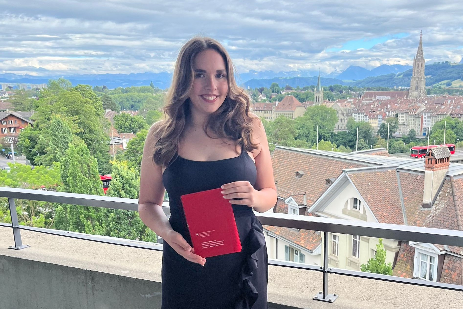 Eine Frau in einem schwarzen Kleid hält auf einem Balkon ein rotes Diplom. Im Hintergrund ist eine malerische Aussicht auf eine Stadtlandschaft mit üppigem Grün, traditionellen Gebäuden und Bergen in der Ferne unter einem bewölkten Himmel zu sehen.