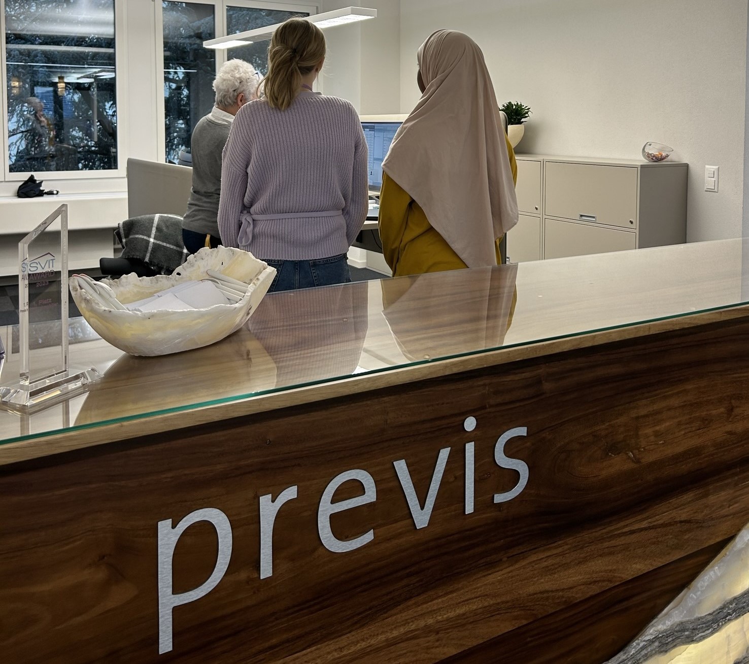 Drei Personen stehen an einem Empfangstresen mit „Previs“-Schild und schauen auf einen Computerbildschirm. Eine Person trägt einen lila Pullover, eine andere ein hellbraunes Kopftuch. Das Büro ist modern, mit grossen Fenstern und Zimmerpflanzen.