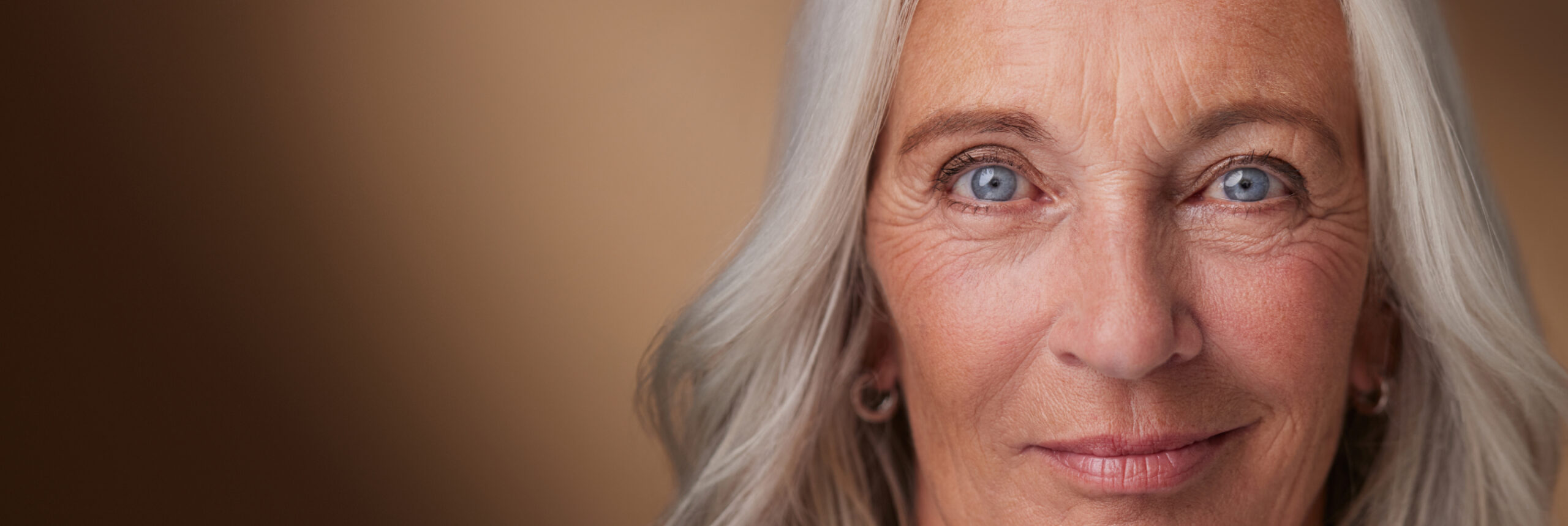 Nahaufnahme einer Frau mit langen Haaren und blauen Augen. Sie hat einen sanften Gesichtsausdruck und blickt vor einem weichen braunen Hintergrund direkt in die Kamera.