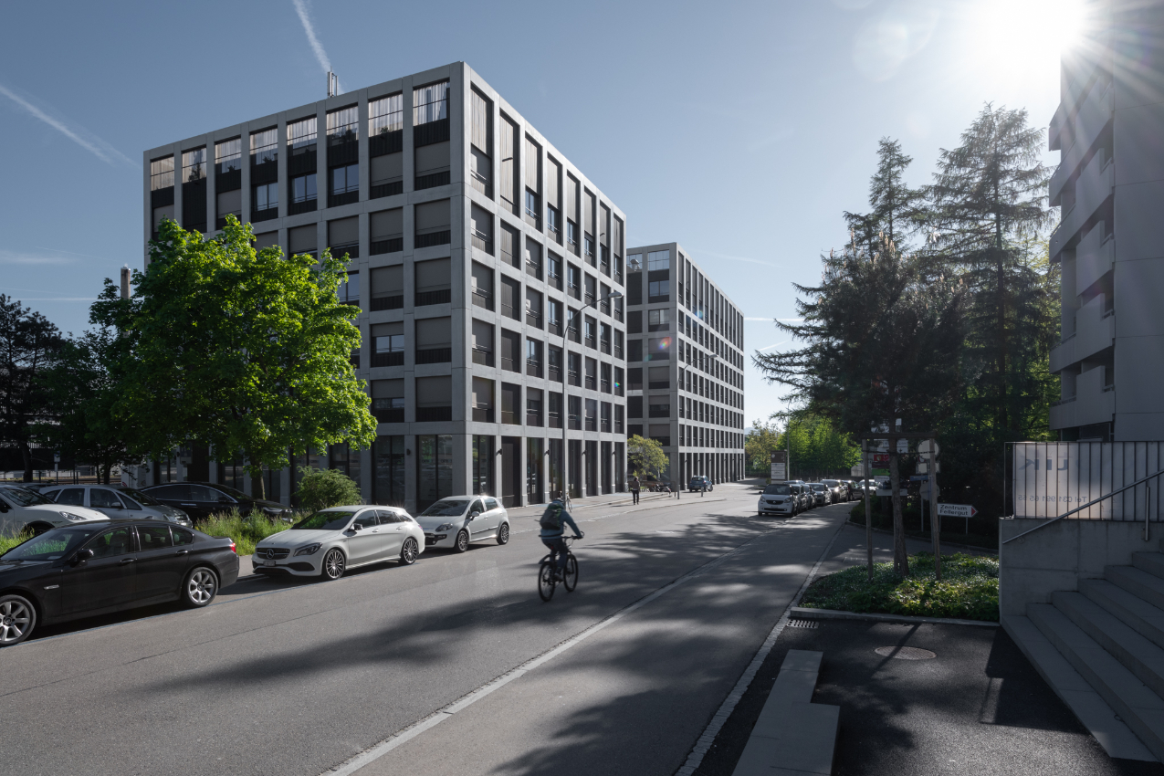 Ein Radfahrer fährt eine sonnige Strasse entlang, die von modernen Bürogebäuden und geparkten Autos gesäumt ist. Bäume und ein strahlend blauer Himmel füllen den Hintergrund, und das Sonnenlicht wirft lange Schatten auf den Bürgersteig.