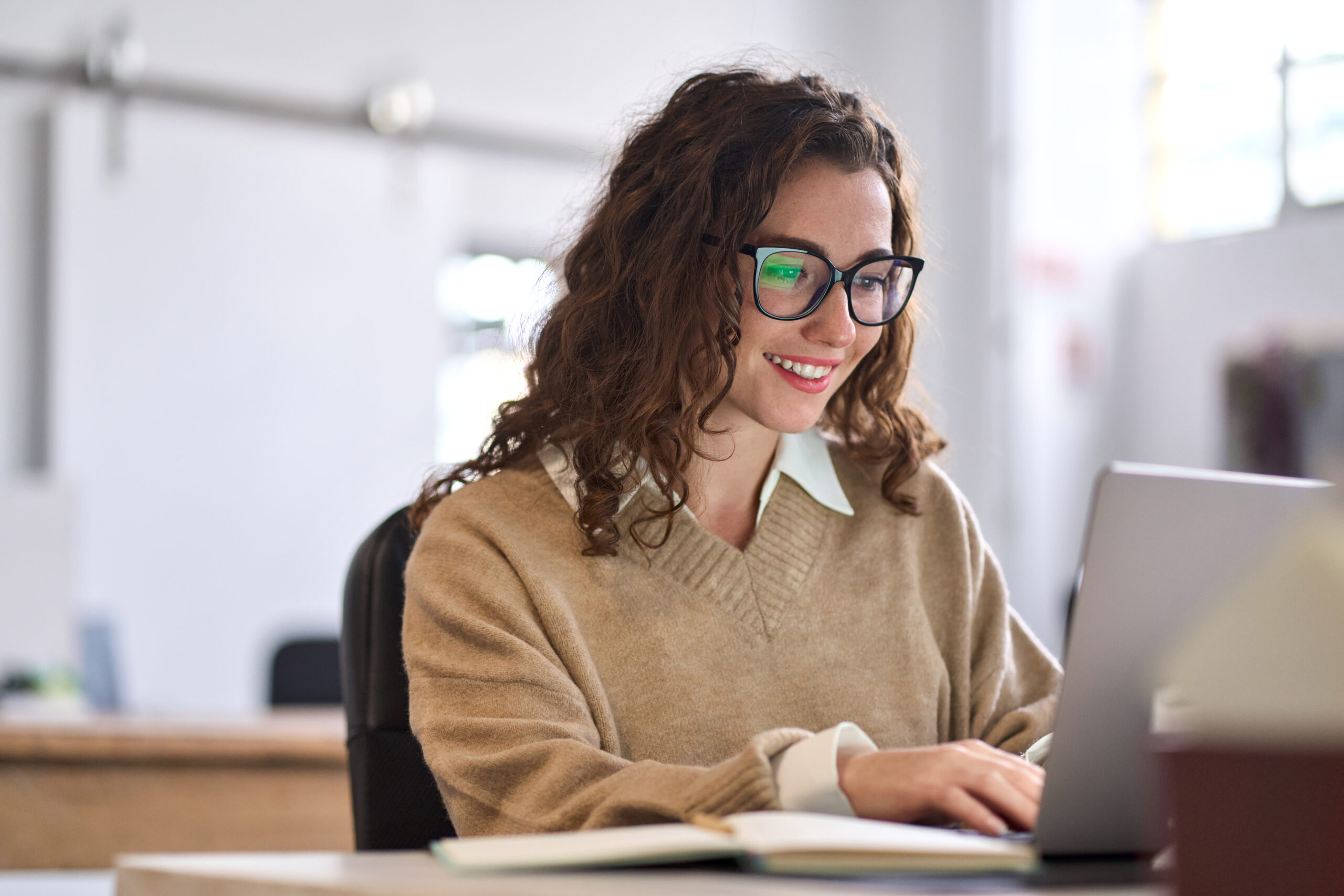 Eine Frau mit lockigem Haar und Brille lächelt, während sie an einem Laptop arbeitet. Sie sitzt in einem lichtdurchfluteten Raum an einem Schreibtisch.