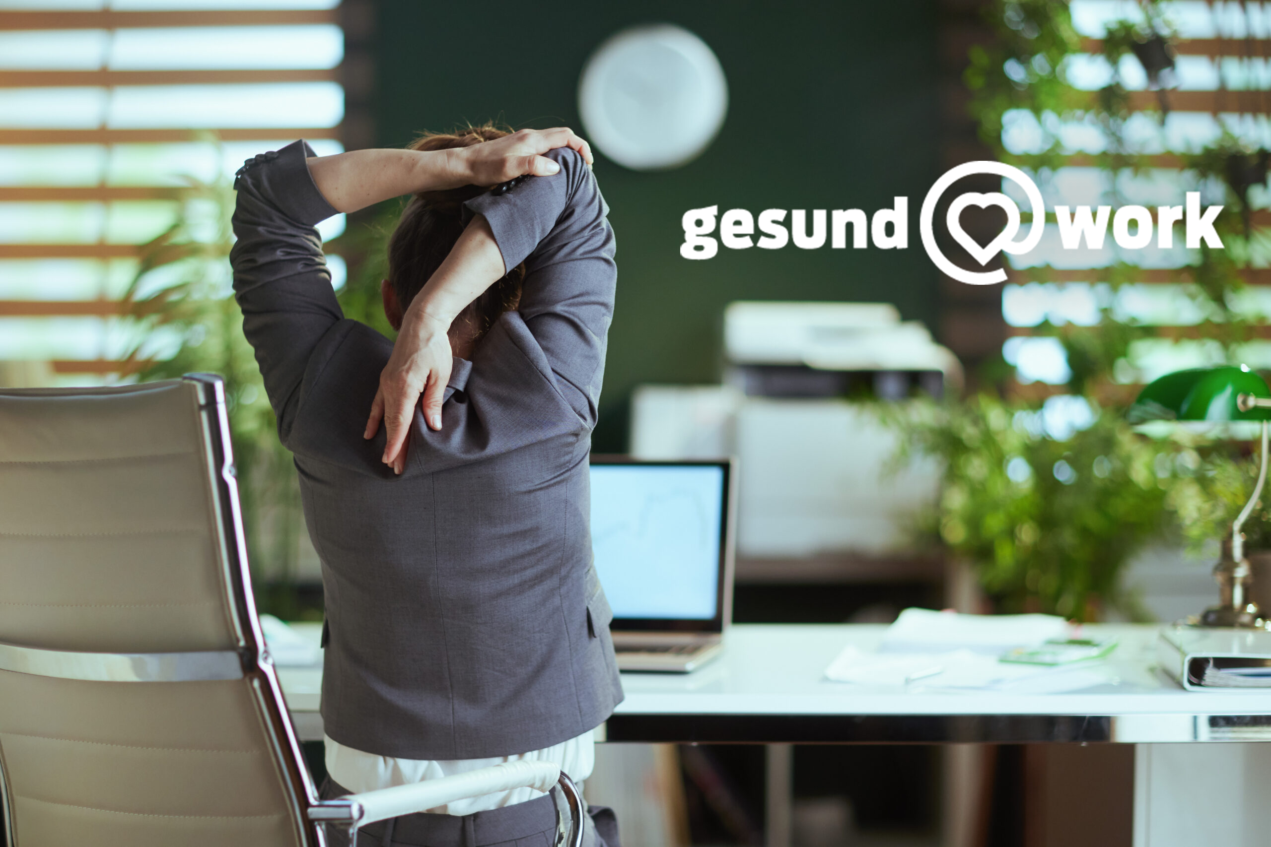 Eine Person im Anzug streckt sich in einem modernen Büro. Sie sitzt auf einem ergonomischen Stuhl, im Hintergrund sind Zimmerpflanzen und ein Computerbildschirm zu sehen. An der Wand prangt der Schriftzug „gesund @ work“.