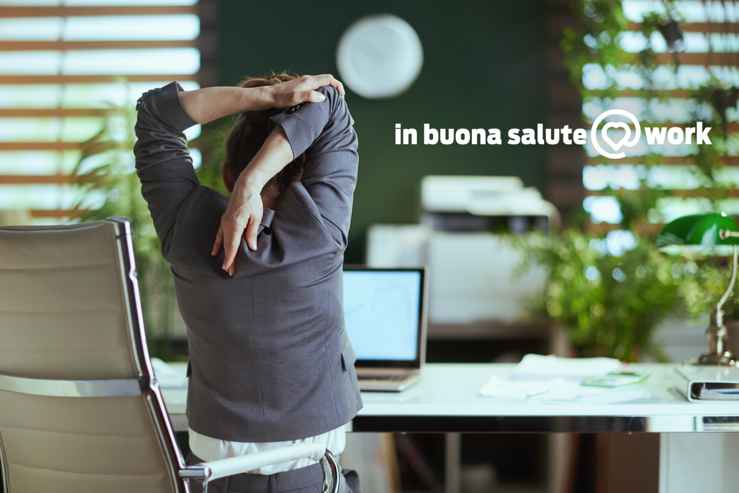 Una persona in giacca e cravatta fa stretching in un ufficio moderno. È seduta su una sedia ergonomica, con piante d'appartamento e lo schermo di un computer sullo sfondo. Sul muro campeggia la scritta “healthy @ work”.