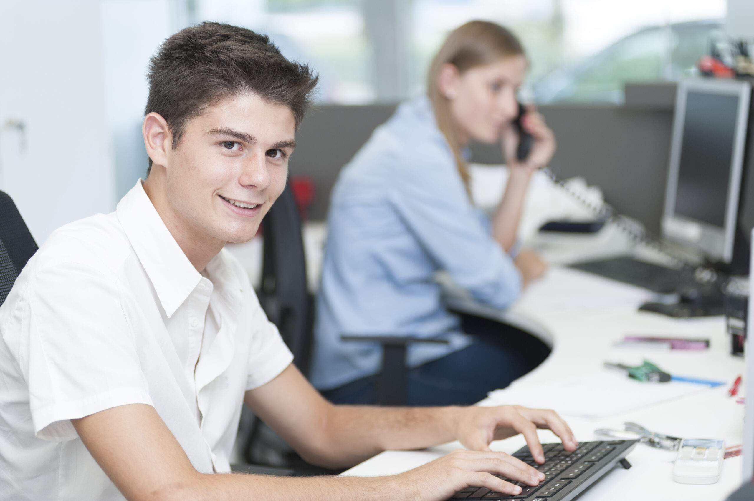 Ein junger Mann im weissen Hemd lächelt in die Kamera, während er in einem Büro auf einer Tastatur tippt. Im Hintergrund telefoniert eine Frau an einem Schreibtisch mit einem Computer.