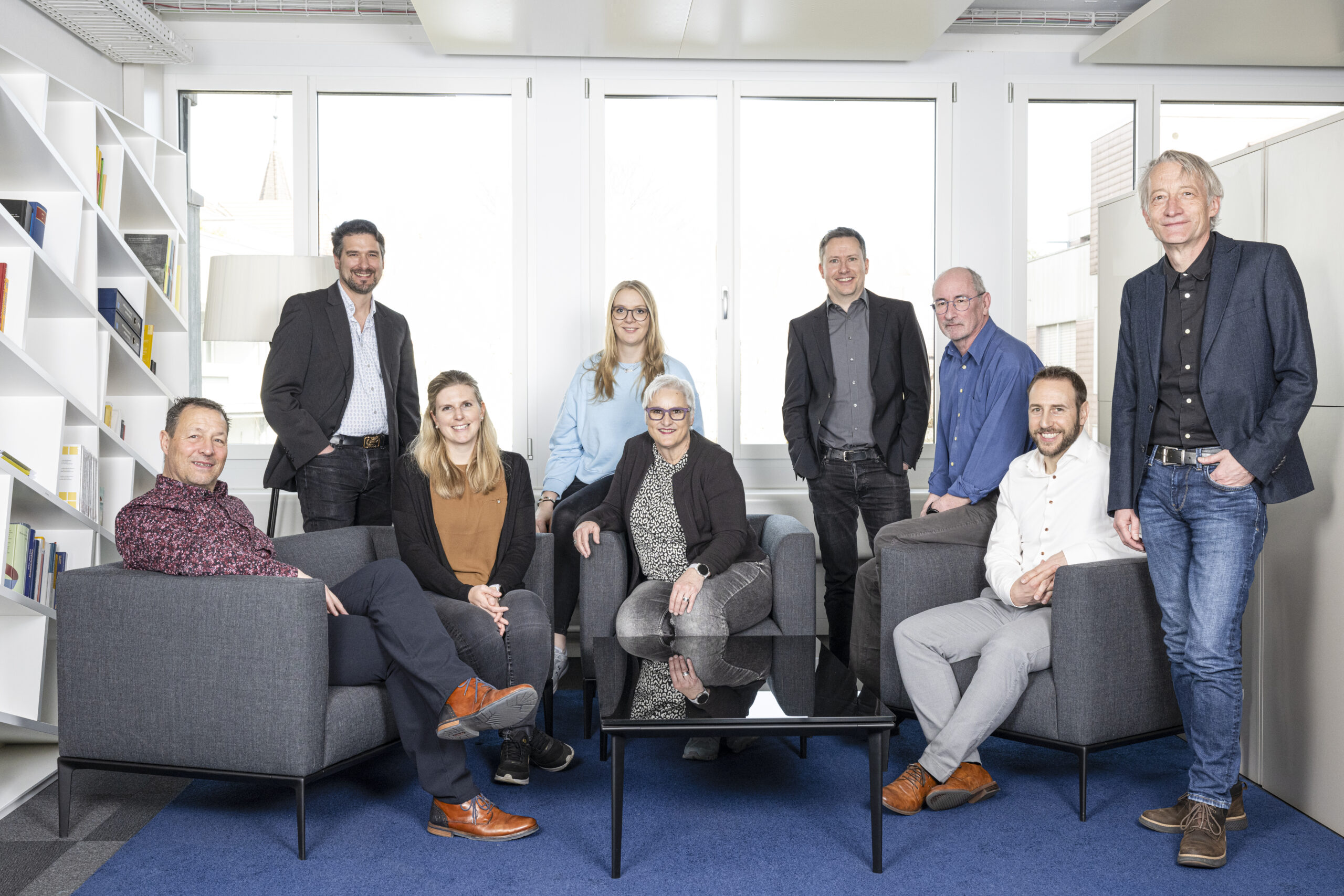 Das Team ICT & Finanzen, bestehend aus drei Frauen und sechs Männern, posiert in einer modernen Büroumgebung mit Bücherregalen und grossen Fenstern. Sie sitzen und stehen um einen Couchtisch aus Glas herum und scheinen zu lächeln.