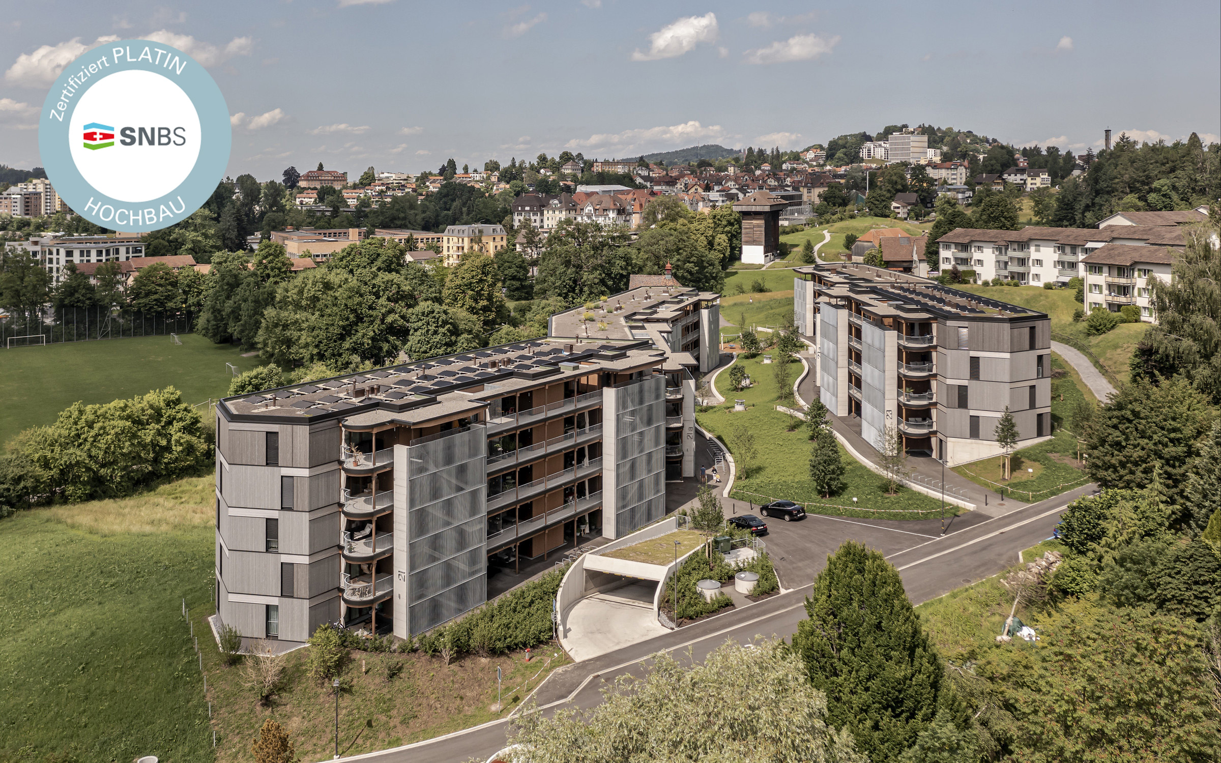 Luftaufnahme moderner Wohngebäude, umgeben von Grünflächen und einer kurvigen Strasse. Die Architektur zeichnet sich durch mehrere Balkone und Sonnenkollektoren auf den Dächern aus. In der oberen linken Ecke ist ein Zertifizierungslogo sichtbar.