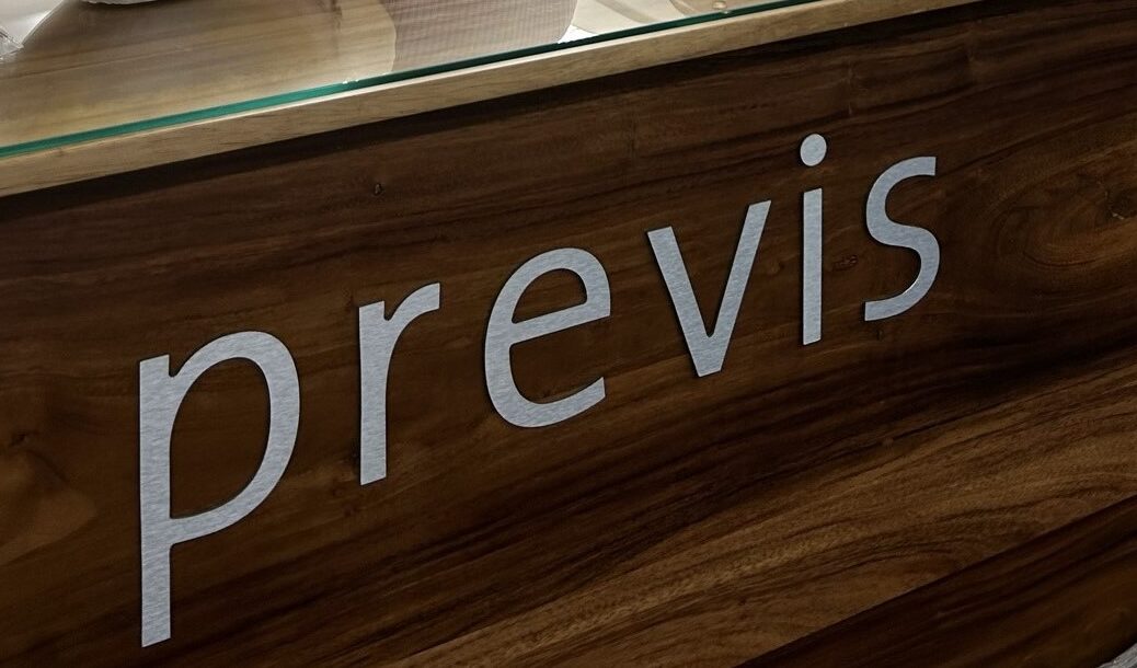 Ein Holzschild mit dem Wort „previs“ in metallischen Buchstaben. Die Oberfläche hat eine satte, dunkle Holzstruktur und die Buchstaben kontrastieren mit ihrem glänzenden, polierten Aussehen.