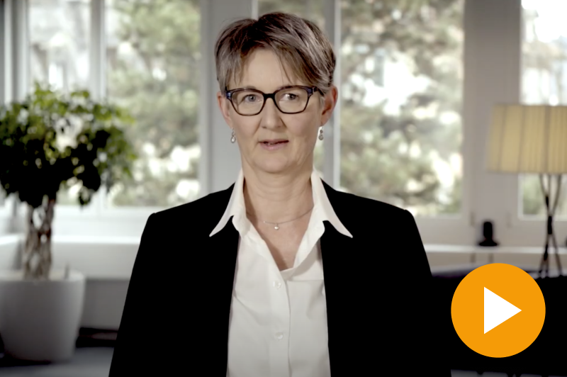 Eine Frau mit kurzen Haaren, Brille, weissem Hemd und schwarzem Blazer spricht in einem gut beleuchteten Raum mit grossen Fenstern und Pflanzen. In der unteren rechten Ecke ist ein orangefarbener Play-Button sichtbar.