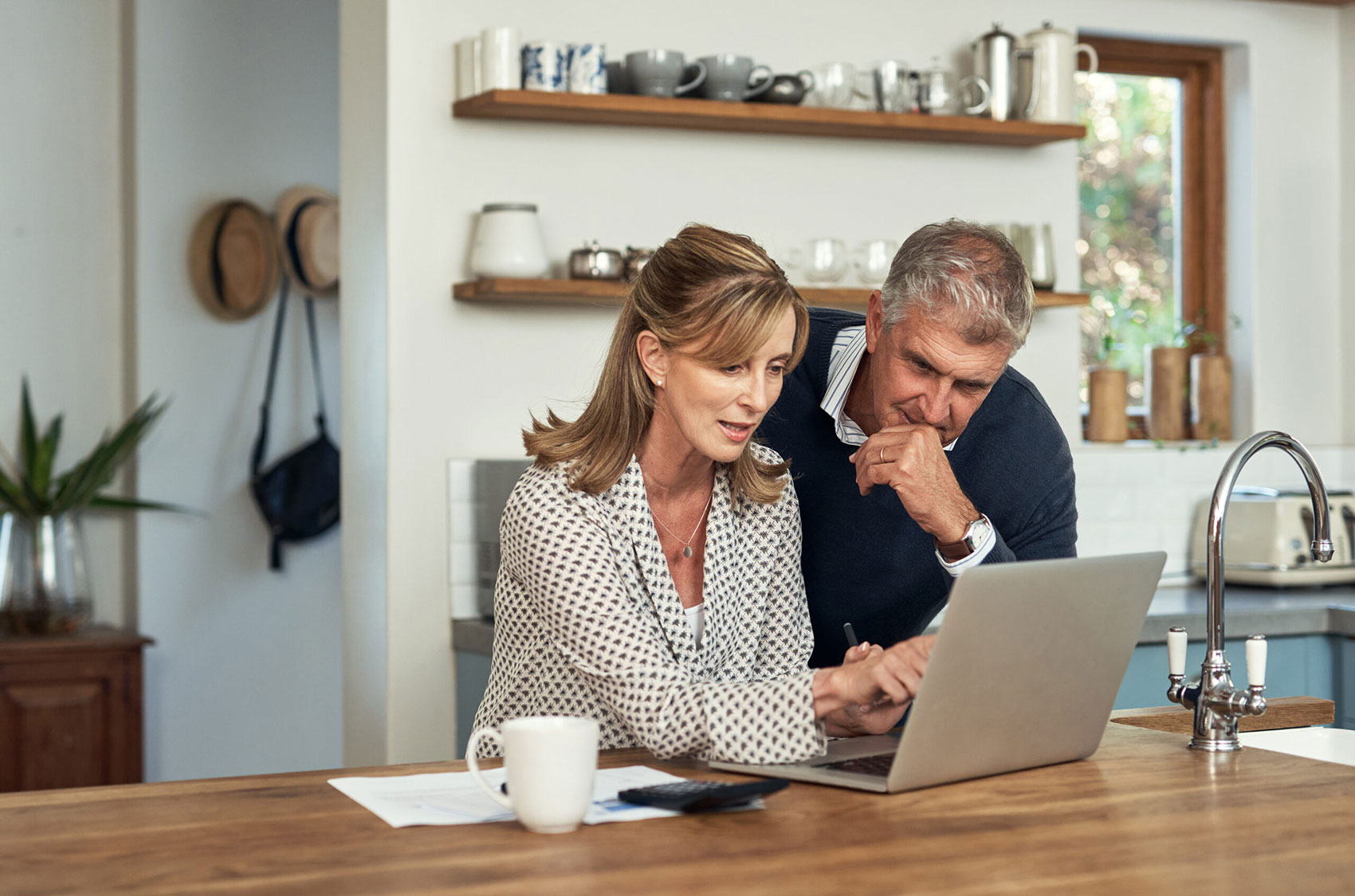 Ein Paar in einer Küche, konzentriert auf einen Laptop. Die Frau, die eine gepunktete Bluse trägt, sitzt, während der Mann, der einen dunklen Pullover trägt, neben ihr steht. Auf der Holztheke vor ihnen liegen ein weisser Becher und Papiere. Dahinter stehen Regale mit Geschirr.